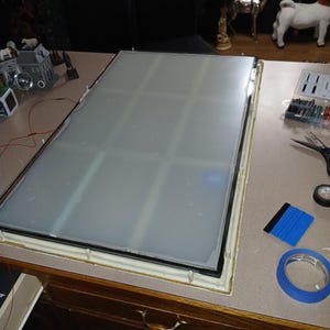 May include: A large, rectangular, frosted glass panel with a black border and a gold-colored frame. The panel is divided into nine sections. Tools and tape are visible on the work surface.