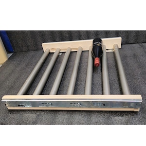 May include: A sliding wine rack featuring light-colored wooden supports and six parallel grey cylindrical rods. A single dark glass wine bottle with a red foil cap rests horizontally between two of the rods. The rack includes metal side rails.