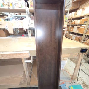 May include: A tall, narrow, dark brown wooden shelf unit. The unit is rectangular with an open front and a simple, clean design. The wood grain is visible, and the finish has a slight sheen. The shelf is empty and appears to be new.