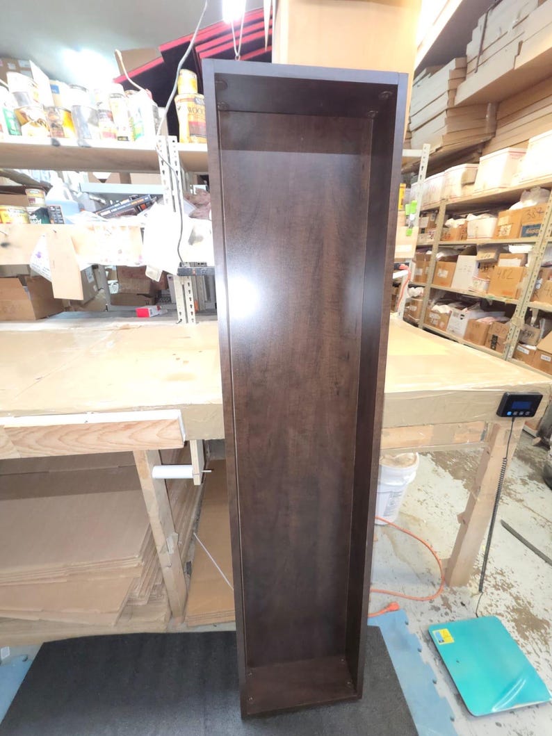 May include: A tall, narrow, dark brown wooden shelf unit. The unit is rectangular with an open front and a simple, clean design. The wood grain is visible, and the finish has a slight sheen. The shelf is empty and appears to be new.
