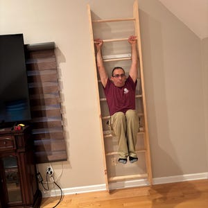 May include: A wooden climbing ladder mounted on a wall. The ladder is made of light-colored wood and has horizontal rungs. A person is hanging from the ladder, with their arms extended above their head.