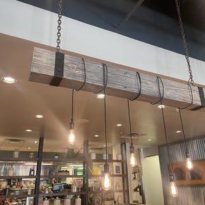 May include: A rustic wooden beam with black metal accents hangs from the ceiling, supporting a row of six pendant lights with exposed black cords and Edison bulbs. The lights are hanging over a kitchen counter with shelves and a gray tiled backsplash.