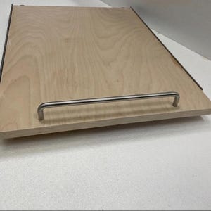 May include: A rectangular wooden tray with a light wood grain finish. It features a stainless steel handle attached to the front edge. The tray has dark edges and is designed for storage or serving.