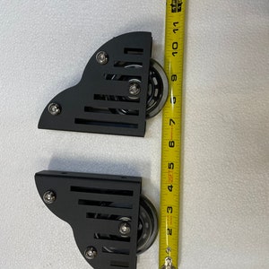 May include: Two black metal caster wheels with clear wheels, each approximately 4 inches in height. The wheels are attached to a bracket with three silver bolts. The image includes a yellow measuring tape.