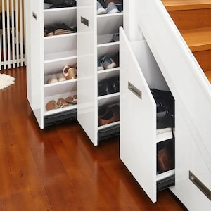 May include: White shoe storage cabinet with multiple shelves, some open and some with pull-out doors. The shelves are filled with various shoes, including sandals, sneakers, and dress shoes. The cabinet is set against a wooden floor.