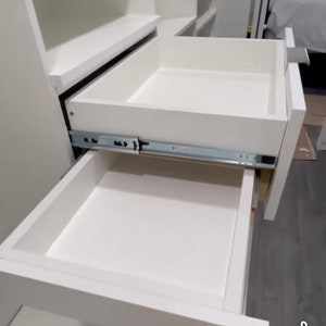 May include: Two white drawers with metal drawer slides are open, revealing the empty interior of the drawers. The drawers are part of a white cabinet.