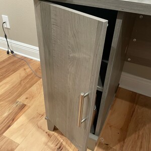 May include: A gray wooden desk with a cabinet door open, revealing shelves inside. The desk has a silver handle and a light wood grain pattern.