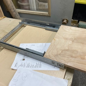 May include: A close-up of a drawer slide system with a piece of wood attached. The slide is made of metal and has a silver finish. The wood is light brown and has a natural grain pattern.