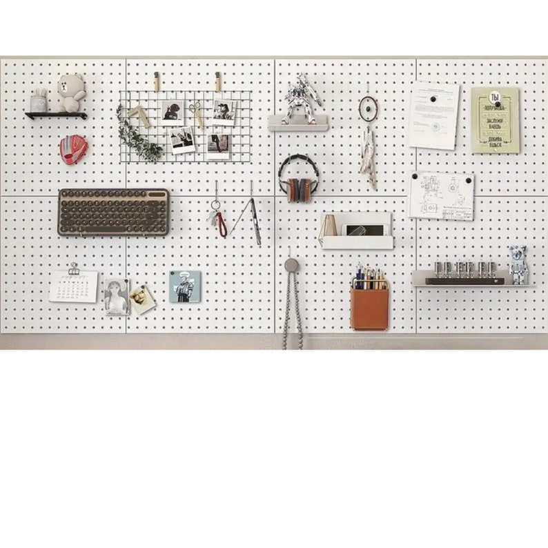 Op de afbeelding: Een wit prikbord met verschillende items eraan bevestigd, waaronder een toetsenbord, een kalender, een draadgaas met foto's, een klein knuffeldier, een koptelefoon, een klein plankje met een bruine leren potloodhouder en een klein plankje met een rij kleine glazen flesjes.