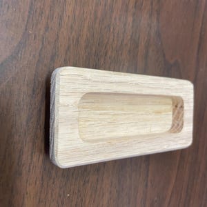 May include: A rectangular wooden drawer pull with a recessed handle. The pull is made of light-colored wood and has a smooth finish.