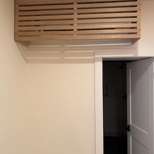May include: A light-colored wooden air conditioner cover featuring a slatted design with horizontal planks. The rectangular enclosure is mounted on the wall above a doorway, concealing the air conditioning unit behind its open-slat structure.