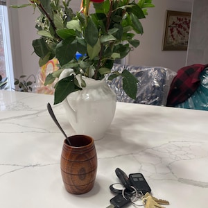 May include: A wooden cup with a dark brown finish and a natural wood grain pattern. The cup is sitting on a white countertop with a set of keys and a silver spoon.