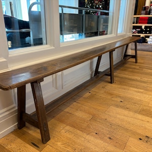 May include: A long wooden bench with a rustic finish and a simple A-frame design. The bench is made of dark brown wood and has a natural, weathered look. It is supported by two sturdy legs on each side.