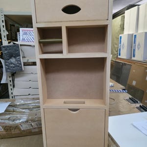 May include: A tall, unfinished wooden storage cabinet with a drawer featuring an oval handle, open shelves, and a lower drawer with a curved handle. The cabinet has a simple, functional design, suitable for various storage needs.
