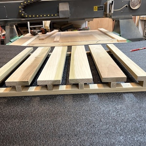 May include: A wooden slatted platform with five slats, each with a T-shaped cut-out at the end. The platform is made of light-colored wood and is sitting on a dark gray surface.