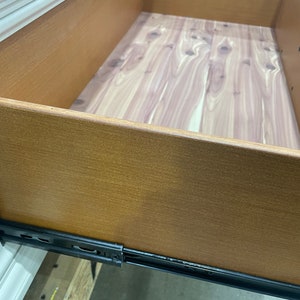 May include: Close-up of a wooden drawer with a cedar-lined bottom. The drawer is open and the wood grain is visible. The drawer is mounted on metal drawer slides.