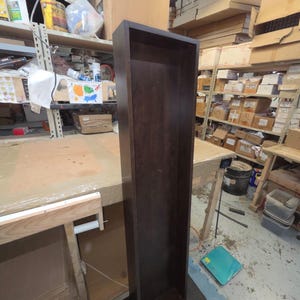 May include: A tall, dark brown wooden shelf unit with a rectangular shape. The unit is empty and stands upright on a gray surface. The shelf is in a workshop setting, with other shelves and tools visible in the background.