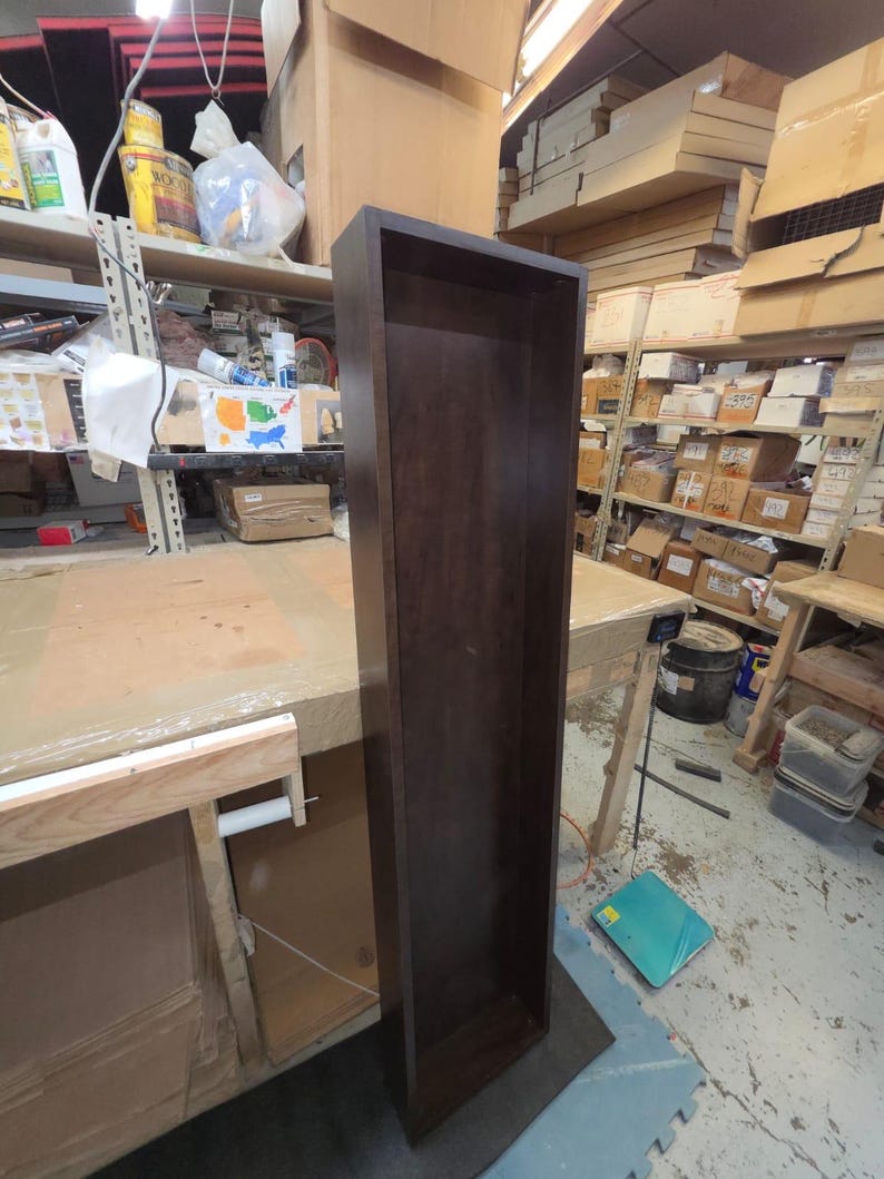 May include: A tall, dark brown wooden shelf unit with a rectangular shape. The unit is empty and stands upright on a gray surface. The shelf is in a workshop setting, with other shelves and tools visible in the background.