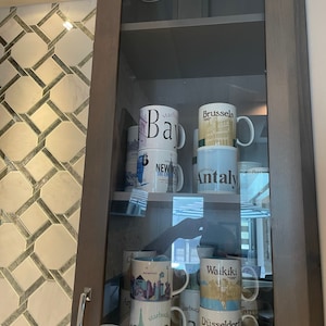 May include: A glass cabinet with shelves filled with various city-themed coffee mugs. The mugs feature images of cities like New York, Bay, Brussels, Antaly, Waikiki, Düsseldorf, and Edinburgh. The mugs are arranged in a staggered pattern on the shelves.