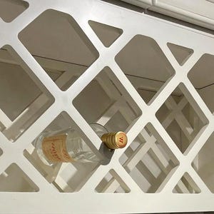 May include: White, geometric wine rack with a diamond pattern. A clear wine bottle with a gold cap is placed inside the rack. The rack is built into a cabinet, suggesting a kitchen or dining area.
