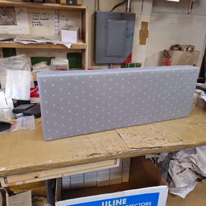 May include: A rectangular, light gray fabric-covered panel featuring a repeating pattern of small, white, evenly spaced dots. The object has a structured, box-like shape with clean, straight edges and a flat front surface, presented in a workshop setting.