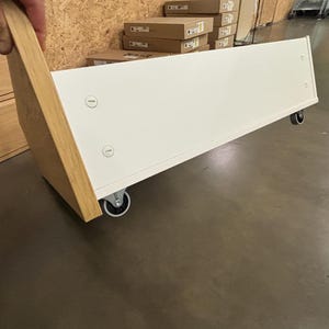 May include: A white cabinet drawer with a light wood side panel, mounted on small caster wheels. The drawer is being held up by a hand. The background includes cardboard boxes and a concrete floor.