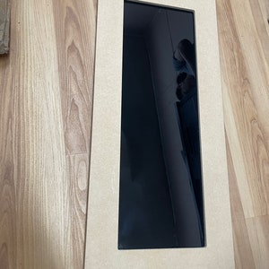 May include: A rectangular, unfinished wooden frame with a black, reflective surface. The frame is made of light brown wood and has a simple, clean design. The black surface appears to be a mirror or a dark panel.