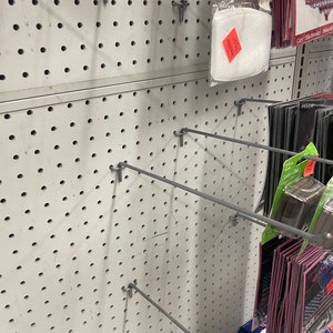 Op de afbeelding: Een witte prikbordwand met verschillende metalen haken en staven. Verschillende producten hangen aan de haken, waaronder een wit luchtfilter, zwart-witte verpakkingen en roze-zwarte verpakkingen.
