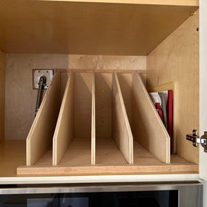 May include: A wooden cabinet organizer with five vertical dividers for storing cutting boards. The organizer is made of light wood and is installed in a cabinet above a stainless steel oven.