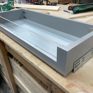 May include: A rectangular, gray wooden drawer with a clear acrylic front panel. The drawer is mounted on metal drawer slides and is designed for installation. The drawer is empty and ready for use.
