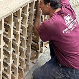 May include: A person is assembling a wooden wine rack. The rack is made of natural wood and has a grid pattern. The person is wearing a maroon t-shirt with the text "www.handyct.com 203-249-7887".
