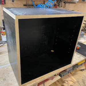 May include: A large, unfinished wooden box with a black interior and light-colored trim. The box is rectangular and appears to be constructed from multiple pieces of wood. The interior is empty, and the box is sitting on a workbench.