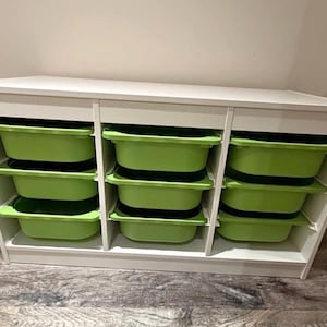 May include: A white storage unit featuring three vertical columns, each containing three removable lime green plastic bins. The rectangular frame is constructed with smooth white panels, providing a structured organization system for various items.