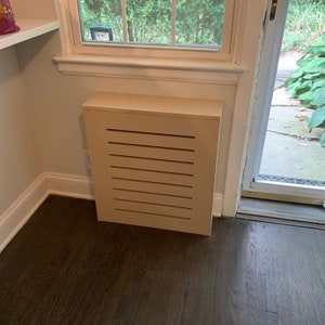 May include: A light brown wooden radiator cover with a slatted design. The cover is installed in a corner of a room with hardwood floors.