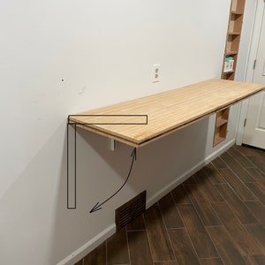 May include: A light-colored wooden wall-mounted folding desk featuring a rectangular surface with a visible wood grain pattern. The desk is attached to a white wall and includes a black line drawing illustrating the folding mechanism and bracket.