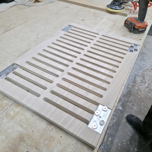 May include: A light-colored wooden panel with evenly spaced horizontal slots, attached with metal brackets. The panel is on a light-colored surface, with tools and a person's leg visible in the background. The panel appears to be part of a woodworking project.
