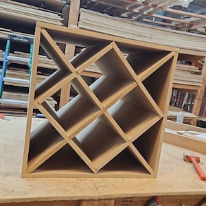 May include: A square wooden wine rack featuring a grid of diagonal dividers that create multiple triangular storage compartments. The rack is constructed from light brown, unfinished composite board with clean, straight edges and a frame.