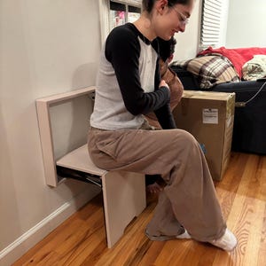May include: A white folding wall-mounted chair with a person sitting on it. The chair is made of wood and has a simple design. The chair is folded up against the wall and is ready to be used.