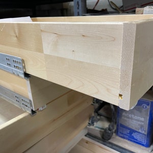 May include: Close-up of unfinished wooden drawer boxes with metal drawer slides. The boxes are made of light-colored wood, showing the natural grain. The drawer slides are attached to the sides, ready for installation. The boxes are stacked.