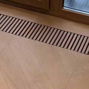 May include: A rectangular, dark brown metal floor vent with parallel slats is installed on a light brown wooden floor. The floorboards are arranged in a herringbone pattern, and a window and door frame are visible in the background.