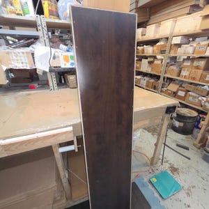 May include: A tall, rectangular wooden panel with a dark brown, wood-grain finish. The panel is standing upright in a workshop setting, with visible screw holes along the edges. The panel is likely a component of a larger piece of furniture.