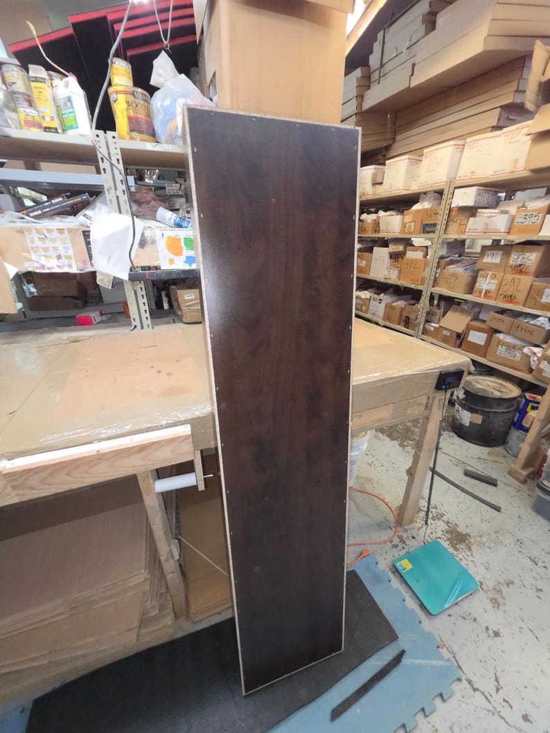 May include: A tall, rectangular wooden panel with a dark brown, wood-grain finish. The panel is standing upright in a workshop setting, with visible screw holes along the edges. The panel is likely a component of a larger piece of furniture.