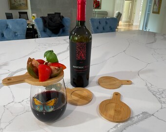 May include: A glass of red wine with a butterfly design on the side. A small wooden cutting board with a handle sits on top of the glass, holding a small salad of tomatoes and basil. A bottle of Apothic Red wine is in the background, along with four wooden coasters.