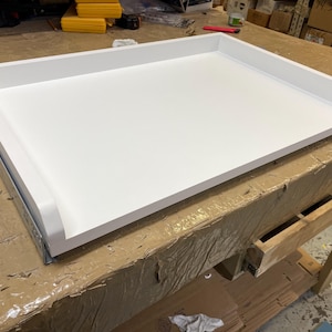 May include: A white rectangular tray with rounded edges sits on a brown surface. The tray is made of a smooth, white material and has a slightly raised lip around the edges.