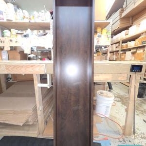 May include: A tall, dark brown wooden cabinet with an open front. The cabinet is rectangular and appears to be unfinished. The wood grain is visible. The cabinet is standing in a workshop setting.