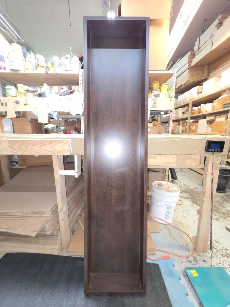 May include: A tall, dark brown wooden cabinet with an open front. The cabinet is rectangular and appears to be unfinished. The wood grain is visible. The cabinet is standing in a workshop setting.