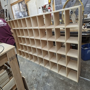 May include: A large, unfinished wooden storage unit with numerous square compartments. The unit is constructed from light-colored plywood and is designed for organizing items. The structure is rectangular and appears to be newly built.