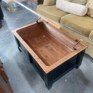 May include: A black wooden storage coffee table with a lift-top lid revealing a cedar-lined interior. The lid is open and the table is sitting on a concrete floor.