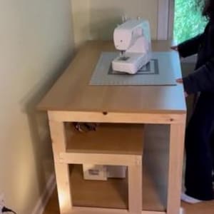 May include: A light-colored wooden sewing table with a white sewing machine on top. The table has a lower shelf and is on wheels. A cutting mat is under the sewing machine. The table is in a room with a window.