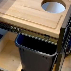 May include: A wooden cutting board with a hole in the center, mounted above a pull-out trash bin. The bin is black and made of plastic.